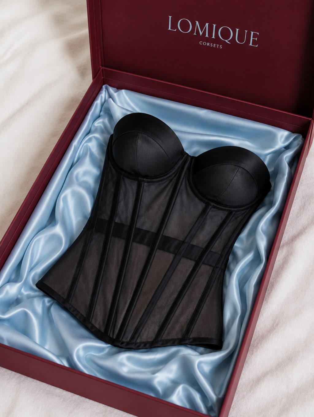 A black sheer mesh and satin underwire corset displayed inside an open dark red LOMIQUE Consoles gift box lined with light blue satin.