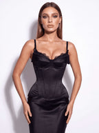 A woman modeling a black satin corset-style dress with lace trim at the bust and thin spaghetti straps, photographed against a plain light background.