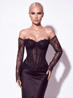 A blonde model wearing a black off-the-shoulder lace corset top with sheer long sleeves and a black satin skirt, posing against a white studio background.